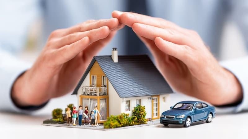 Hands sheltering a model home, car, and family figures, representing protection